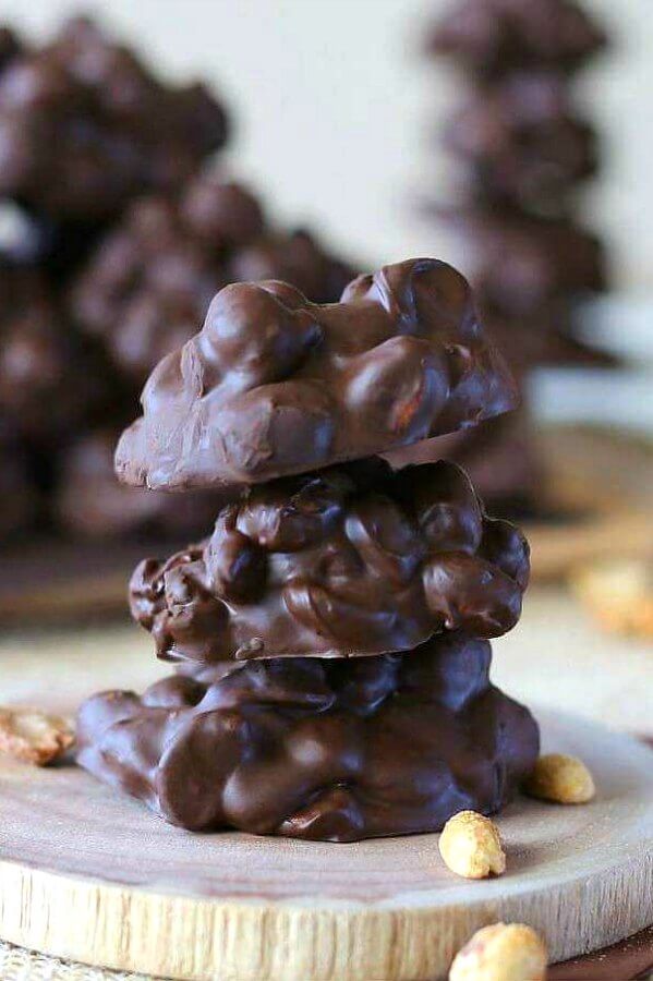 Slow Cooker Chocolate Clusters are glistening in a stack of four candies. Sitting on a wooden coaster with peanuts sprinkles around.