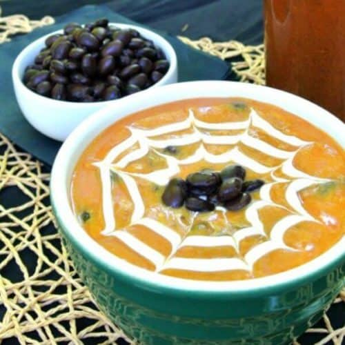 Spicy Pumpkin Black Bean Soup is in a green bowl and has a cream spider web drawn in the top. Black beans are dotted in the center. Great Halloween visual.