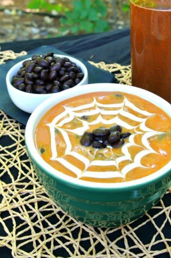 Spicy Pumpkin Black Bean Soup is in a green bowl and has a cream spider web drawn in the top. Black beans are dotted in the center. Great Halloween visual.