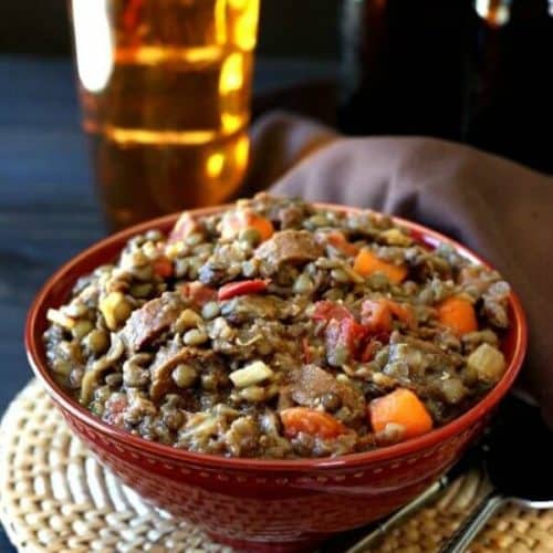Vegan Lentil Sausage Casserole is piled high in a red porcelain bowl and sitting on a woven mat. A gold glass filled with beer sits behind.