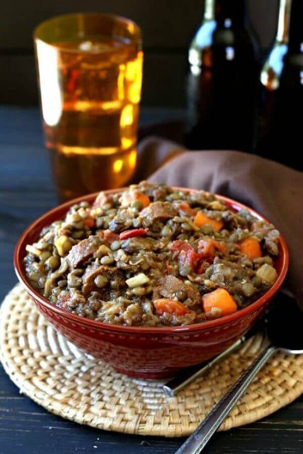 Vegan Lentil Sausage Casserole is piled high in a red porcelain bowl and sitting on a woven mat. A gold glass filled with beer sits behind.
