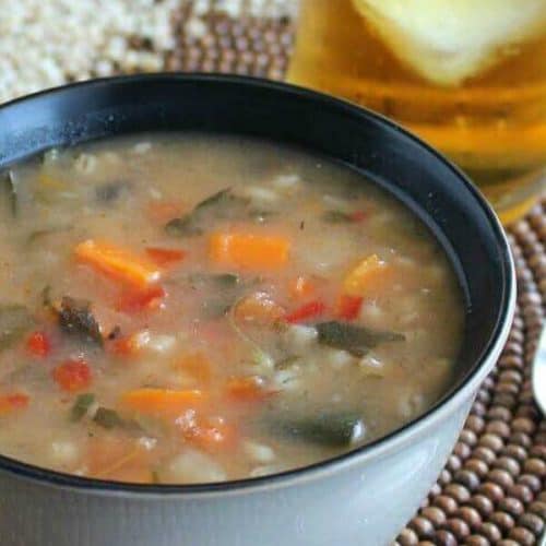 Vegetable Barley Soup is a side view of the rich brothy soup showing carrots, barley, tomatoes, spinach, and more. Beige bowl with a black interior on a dark brown wooden beaded mat.