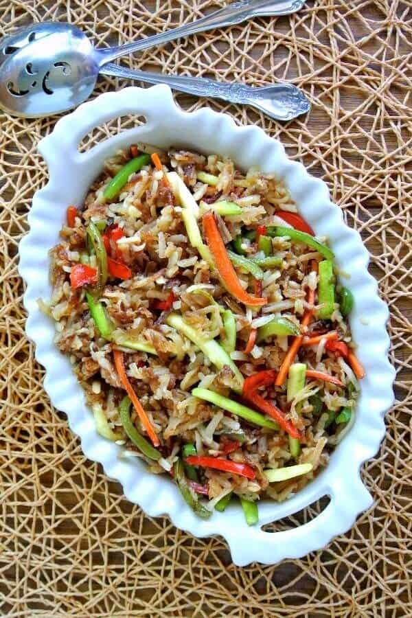 Rainbow Veggie Hashbrown Potatoes are fried together with carrots, bells, zucchini and onions. An overhead photo and piled high in a green and white scalloped bowl on an open-weave beige mat. Serving spoons on the side.