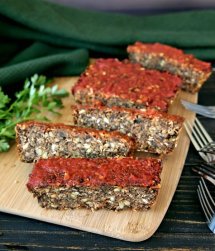 Vegan Meatloaf with Chickpeas, Green Split Peas and Mushrooms is sitting on a breadboard and tilted forward with half of the loaf sliced. It's also topped with marinara sauce. A teal cloth is behind the loaf.