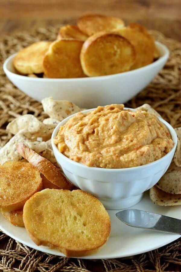 Artichoke Chipotle Hummus is golden in color and is heaped in a white bowl. It's sitting on a white plate with crostini toasts sitting around.