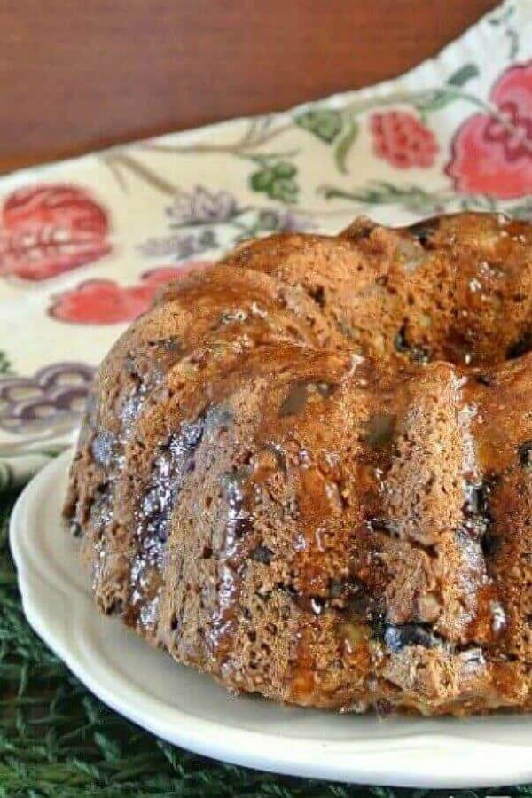 Bundt Cake Fruitcake is sitting prettily on a white scalloped plate. The glistening glaze is dripping over the top with pieces of tiny sweet fruit pieces showing throughout the sides.