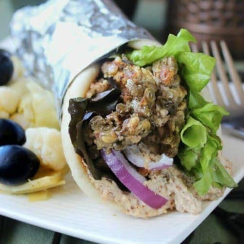 Lentil Panini Burgers are rolled into a pita flatbread and are sitting on a white square plate with olives, onions and lettuce peeking out.