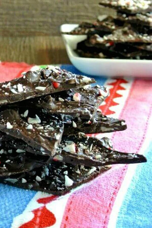 chocolate Peppermint Bark is stacked 7 chunks high. All are tilted and balanced with chips of peppermint on each top. Sitting on a blue, white, raspberry and red cloth.