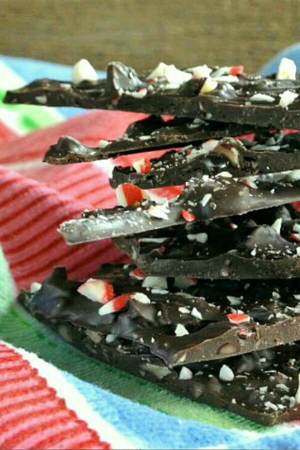 chocolate Peppermint Bark is stacked 7 chunks high. All are tilted and balanced with chips of peppermint on each top. Sitting on a blue, white, raspberry and red cloth.