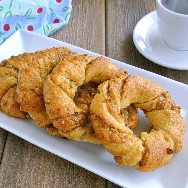Crescent Roll Breakfast Rings are sprawled across a rectangle white plate on a rough wood table top. 5 Rings are overlaying each other and glistening with sweetness.