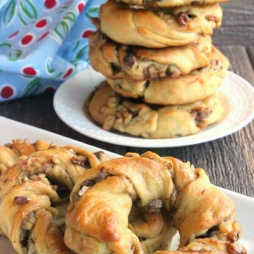 Crescent Roll Breakfast Rings are sprawled across a rectangle white plate on a rough wood table top. A stack of 5 more are stacked high on a plate behind.