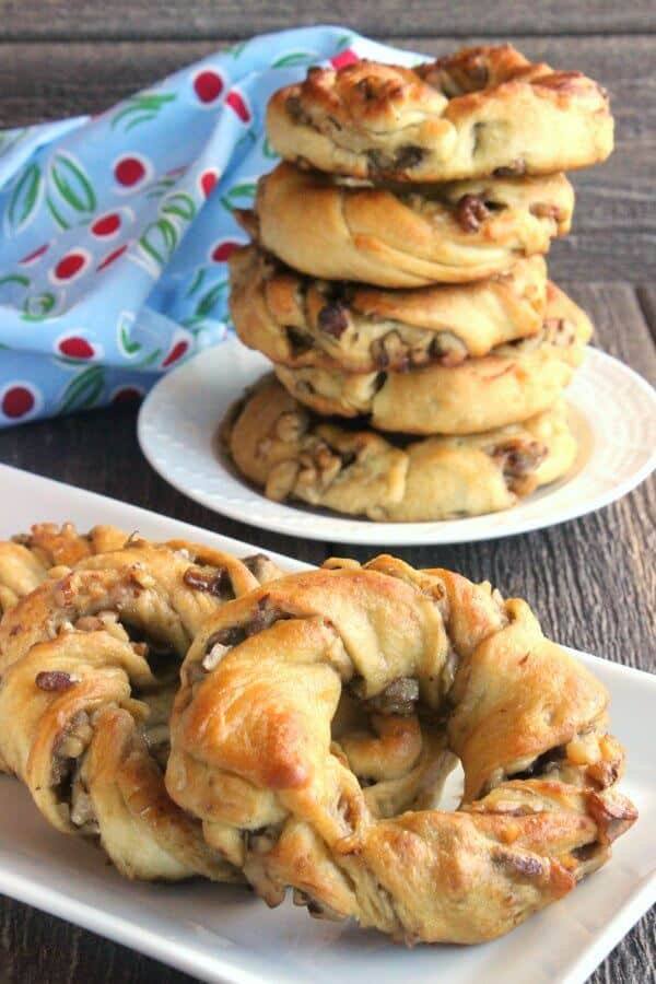 Crescent Roll Breakfast Rings are sprawled across a rectangle white plate on a rough wood table top. A stack of 5 more are stacked high on a plate behind.