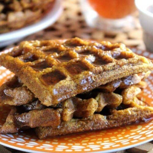 Vegan Pumpkin Waffles are cut into triangles and stacked high on an orange and white patterned plate. Maple syrup is filling the crannies and overflowing intro the plate.