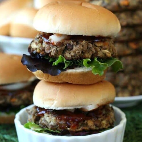 Lentil Burger Sliders are stacked two high in a white scalloped dish. All are sitting on a green mat. Lots of condiments peeking out of the sliders.