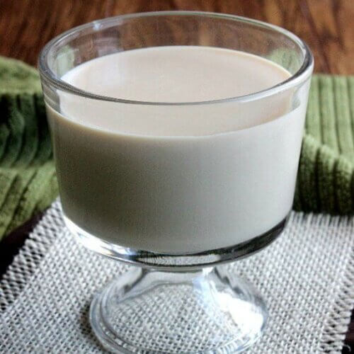 Slow Cooker Sweetened Condensed Milk is in a footed glass and is standing on a white burlap square and a chocolate colored napkin then it's on a olive green cloth.