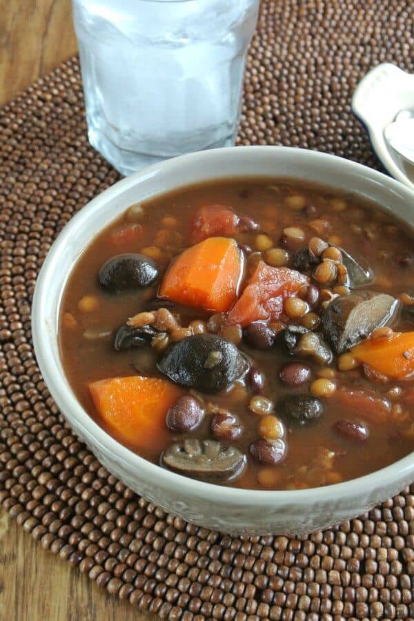 A rich dark soup is filling a gray bowl and show a rich brown broth with carrots, lentils, mushrooms, barley, tomatoes and more. On a brown beaded mat.