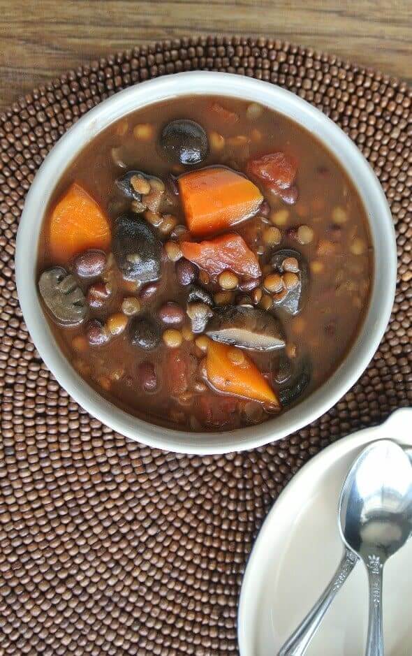 Slow Cooker Lentil Vegetable Soup is an overhead view and is filling a gray bowl and show a rich brown broth with carrots, lentils, mushrooms, barley, tomatoes and more. On a brown beaded mat.s