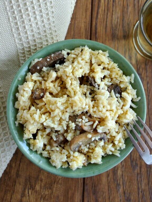 Healthy Slow Cooker Mushrooms and Rice is photographed overhead and is served in a green pottery bowl with contrasting mushrooms sticking out of the rice. Ivory cloth behind and a glass of chardonnay.
