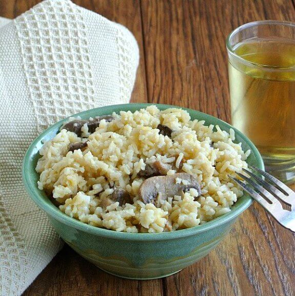 Healthy Slow Cooker Mushrooms and Rice is served in a green pottery bowl with contrasting mushrooms sticking out of the rice. Titled forward with a fork leaning on the rim and a glass of chardonnay behind.