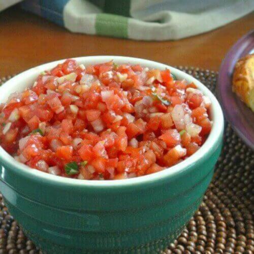 Tomato Bruschetta is a gorgeous red color in a small white rimmed green bowl. Sitting on a chocolate colored beaded mat with golden crostini toasts to the side.