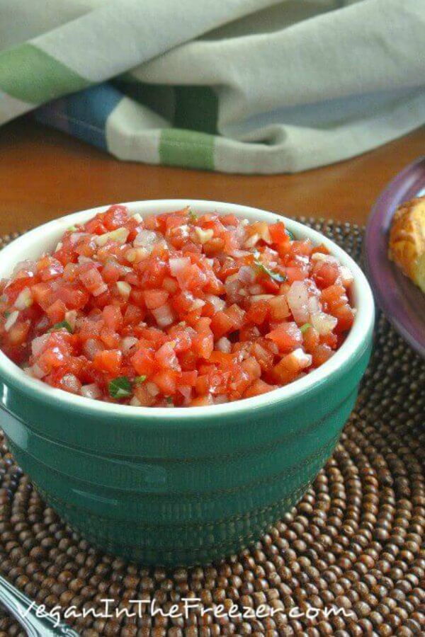 Tomato Bruschetta is a gorgeous red color in a small white rimmed green bowl. Sitting on a chocolate colored beaded mat with golden crostini toasts to the side.