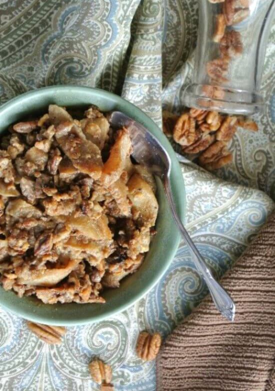 Slow Cooker Apples and Oats is an overhead photo with the cooked brunch in a green bowl sitting on a blue and green paisley cloth. Pecans sprinkled around.