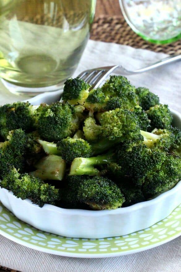 Healthy Balsamic Roasted Broccoli is piles high in a whie scalloped serving dish and that is sitting on a green and white printed plate. A fork is piercing a piece and holding it up.