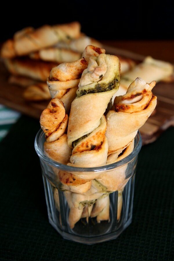 Healthy Crescent Roll Pesto Appetizers are standing up ton end in a wide mouth clear drinking glass. A stack of the remaining finger food are on a cutting board behind.