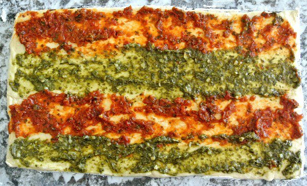 Healthy Crescent Roll Pesto Appetizers has one layer of the crescent roll pressed out on a granite cutting board. The two pesto varieties are spread in four stripes across the dough. Waiting to be covered with the other crescent roll layer.