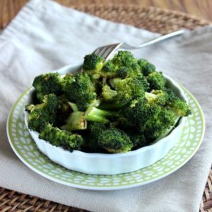 Balsamic Roasted Broccoli is piled high in a white scalloped serving dish and that is sitting on a green and white printed plate. A beige cloth napkin is below all.