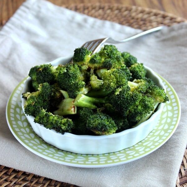 Healthy Balsamic Roasted Broccoli is piled high in a white scalloped serving dish and that is sitting on a green and white printed plate. A beige cloth napkin is below all.