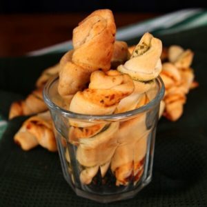 Healthy Crescent Roll Pesto Appetizers are standing up on end in a wide mouth clear drinking glass. A stack of the remaining twists are on a cutting board behind.