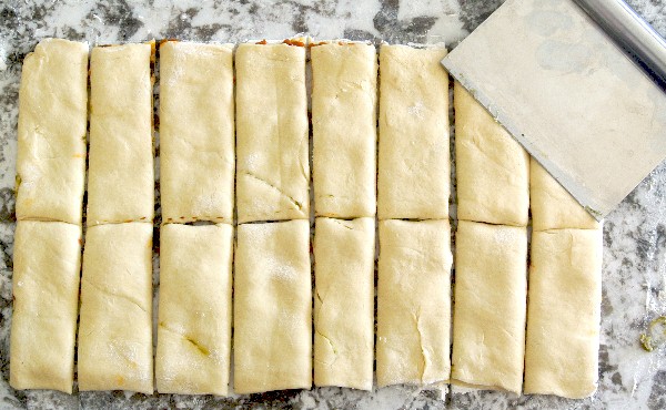 Healthy Crescent Roll Pesto Appetizers are laying on a granite cutting board. The two crescent roll layers are done and a pastry cutter has them sliced into perfect rectangles.