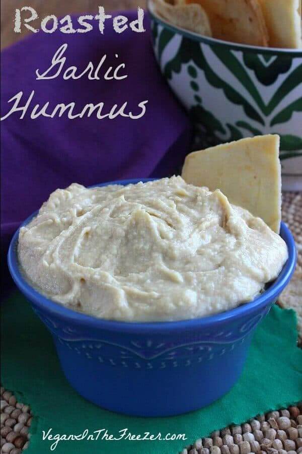 Roasted Garlic Hummus is piled high in a blue bowl set against green and purple.