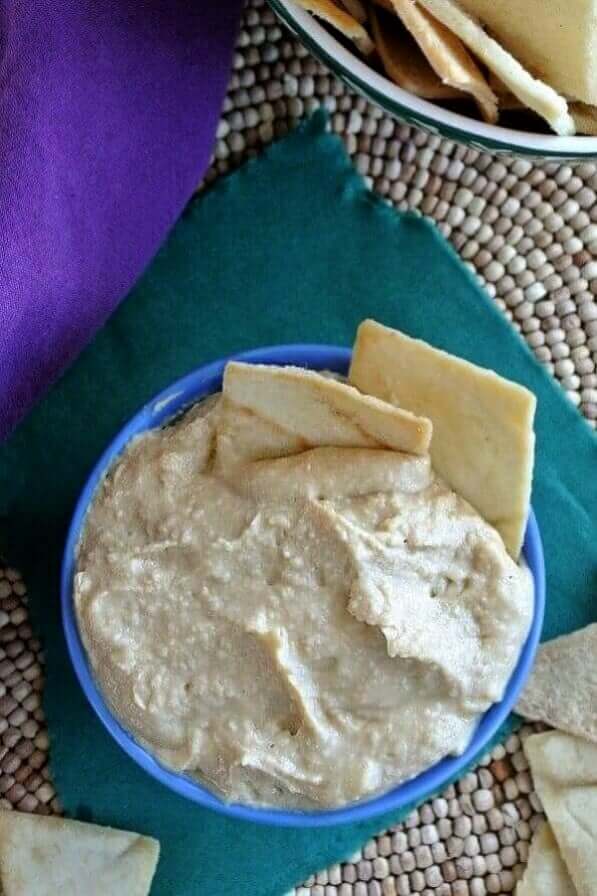 Roasted Garlic Hummus is in a blue bowl and the photo is taken from above. Set against green and purple.