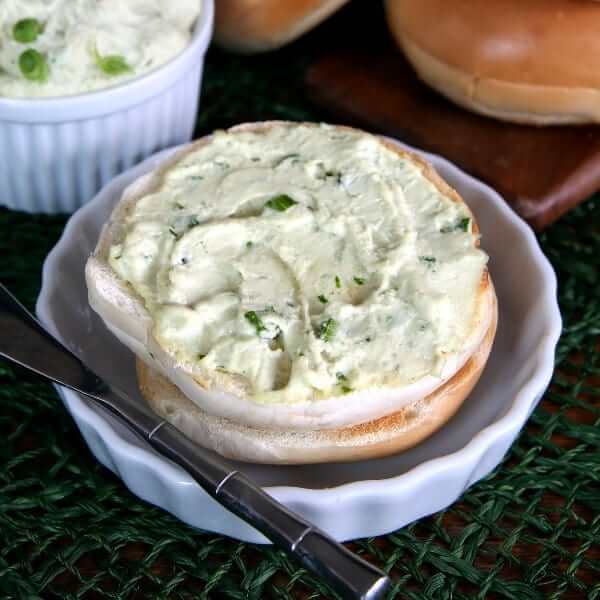 Dairy Free Cream Cheese is spread on a toasted bagel half with chives peeking through and a spreading knife on the side.