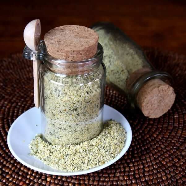 Nut Free Vegan Parmesan Cheese is in a cork topped jar with some spilled on the plate it's sitting on.a tipped corked jar behind.