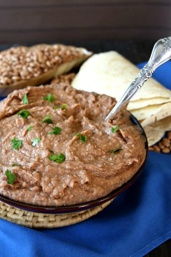 Slow Cooker Refried Beans are piled in a blue bowl with a spoon waiting to dish them up. Tortillas on the side.