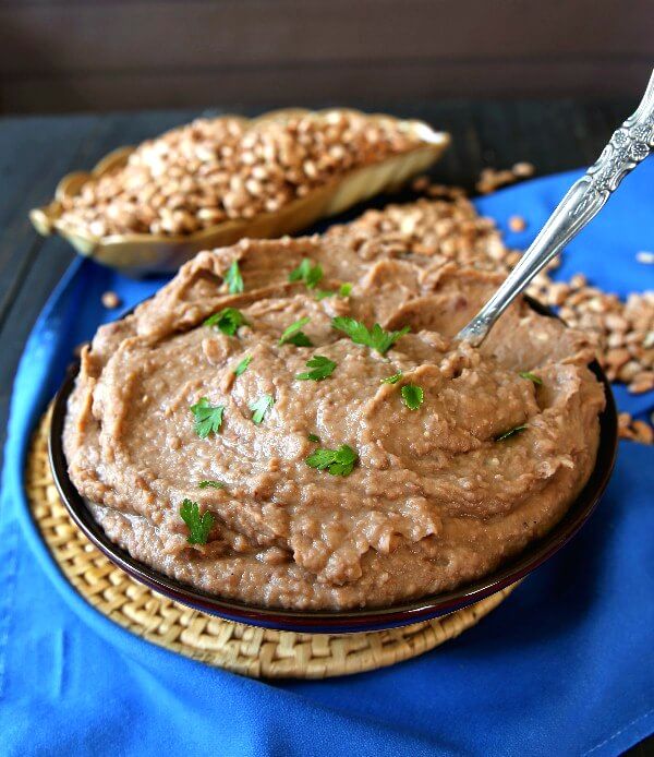 Slow Cooker Refried Beans are piled in a blue bowl with a spoon waiting to dish them up. Blue cloth underneath.