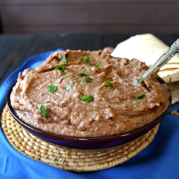 Slow Cooker Refried Beans are piled in a blue bowl with a spooon waiting to dish them up.