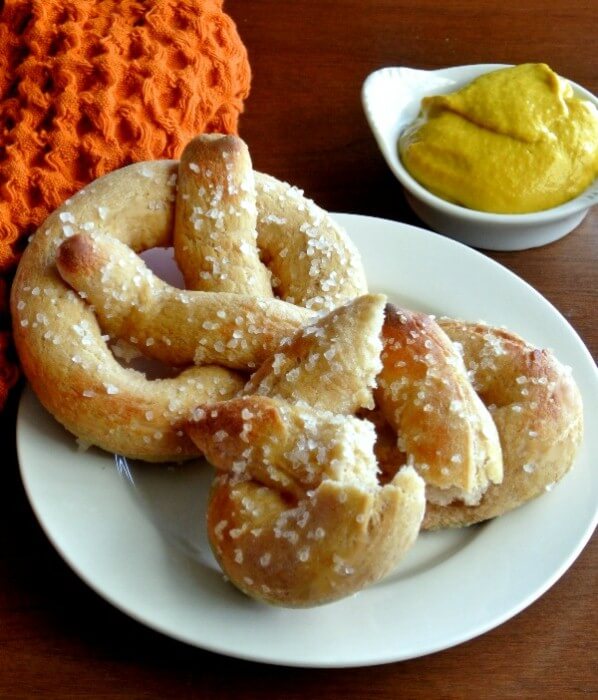 Two homemade soft pretzels are sitting on a small white plate and one is torn in half. Mustard is on the side.