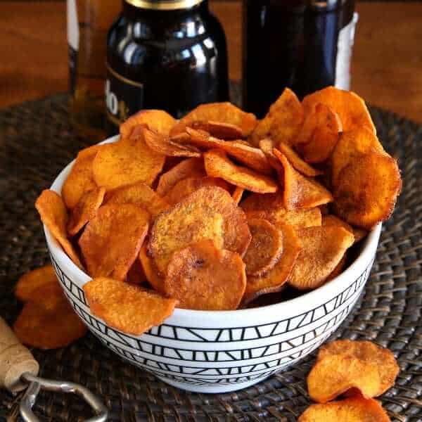 Air Fryer Sweet Potato Chips are overflowing a geometric black and white bowl tilted forward and spilling onto the brown beaded mat.