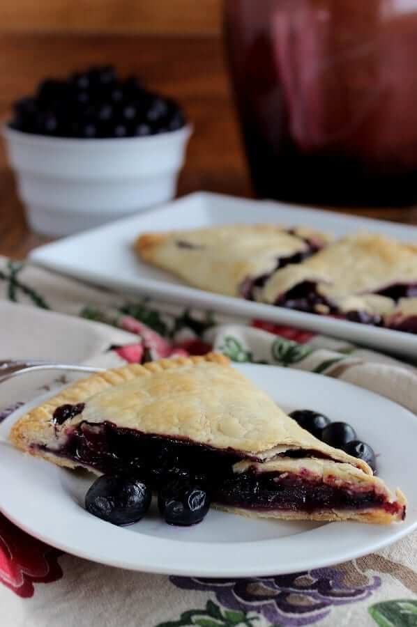 Side view of a slice of blueberry pie on a white plate and a fruit printed hand towel with the remaining slices behind.