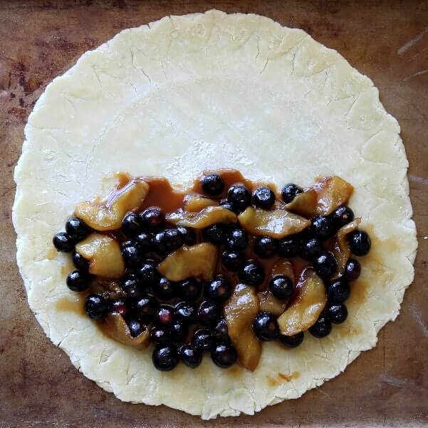 Overhead view of a raw round pie crust with the cooked filling of blueberries and apples on half.