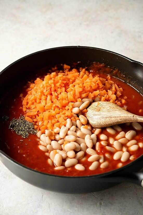 A tilted skillet with fresh ingredients being added to Tuscan Pasta and Beans.