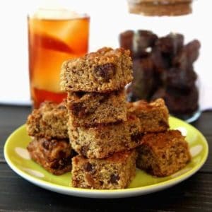 Stack and pile of banana bread squares infont of a glass of iced tea and a jar of dates.