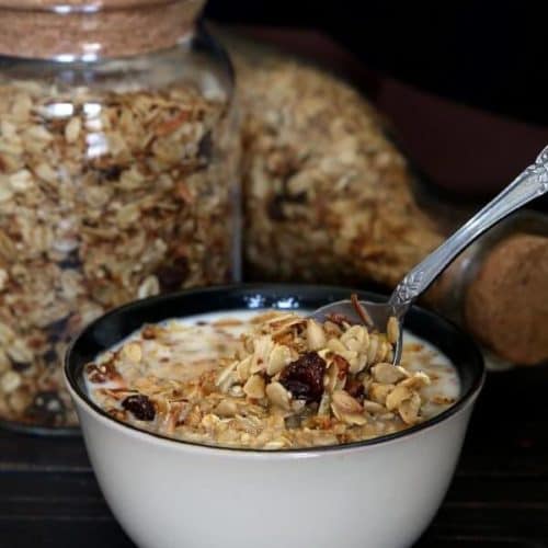 A bowl full of nut free granola is being scooped up for a bite. With two jars of homemade cereal behind.