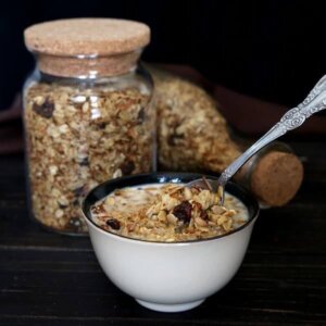 A bowl full of nut free granola is being scooped up for a bite. With two jars of homemade cereal behind one flipped on it's side.