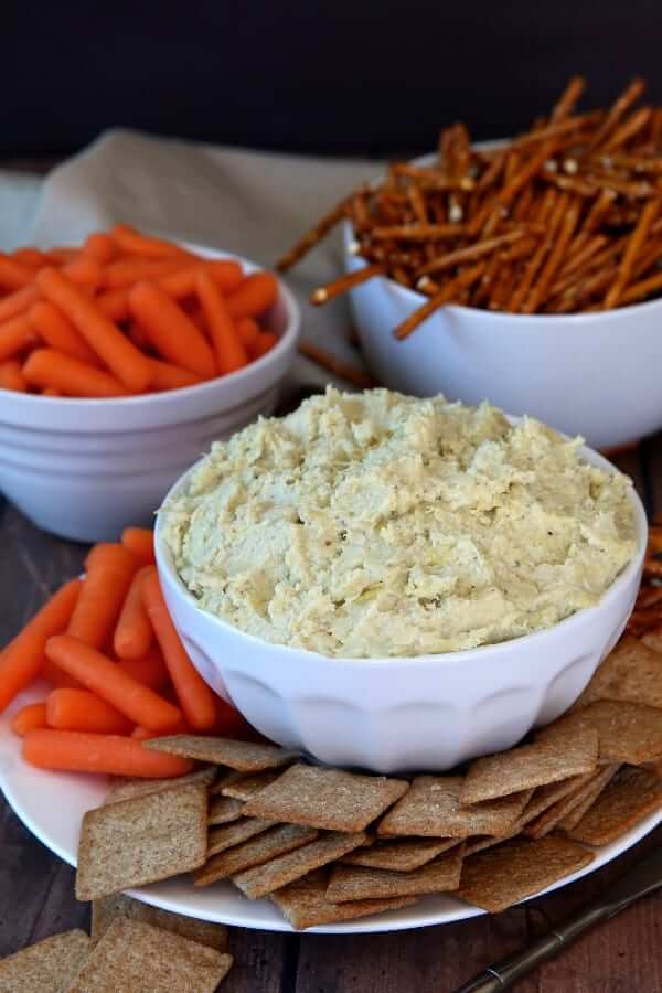 White bowl filled with artichoke hearts dip with wheat thins and baby carrots piled around the bowl.