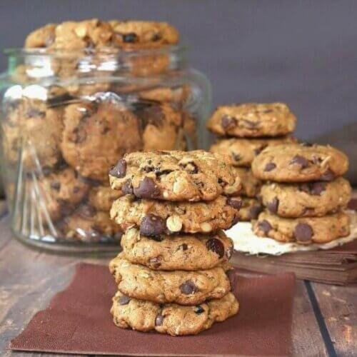 Five cookies are stacked high on a brown napkin with chocolate chips pied in a cookie jar behind.
